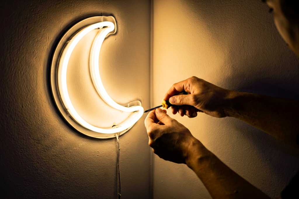 Close-up of hands working with a glowing warm white neon sign in a dim room